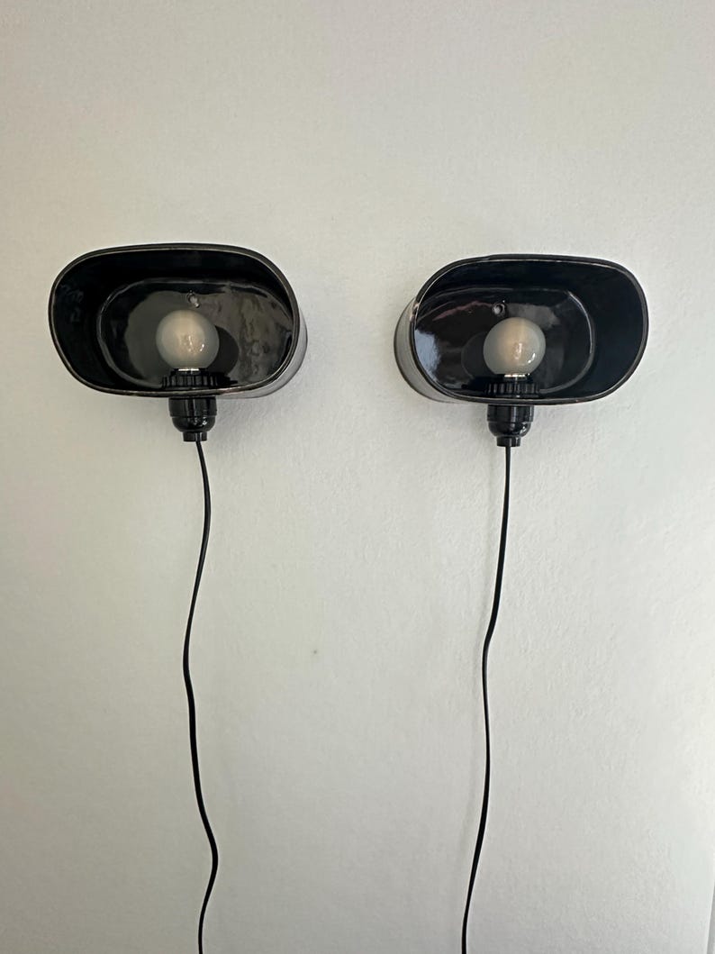 May include: Two black, oval wall sconces with visible light bulbs. Each sconce has a black cord and is mounted on a white wall. The sconces have a minimalist design, with a simple, industrial aesthetic.