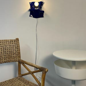 May include: A wall-mounted lamp featuring a glossy, deep blue, undulating ceramic shade with a single exposed light bulb. The lamp is connected to a thin white power cord that hangs vertically down the wall.