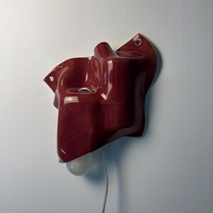Puede incluir: Un aplique de pared de cer&aacute;mica en color rojo oscuro brillante con una forma org&aacute;nica, irregular y ondulada. La pieza cuenta con dos peque&ntilde;os orificios de montaje circulares con ojales met&aacute;licos y una bombilla blanca que sobresale.