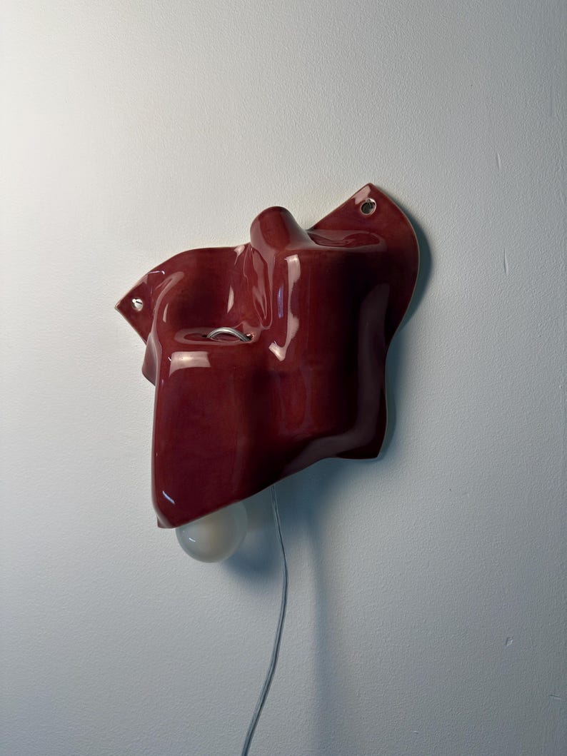 Puede incluir: Un aplique de pared de cer&aacute;mica en color rojo oscuro brillante con una forma org&aacute;nica, irregular y ondulada. La pieza cuenta con dos peque&ntilde;os orificios de montaje circulares con ojales met&aacute;licos y una bombilla blanca que sobresale.