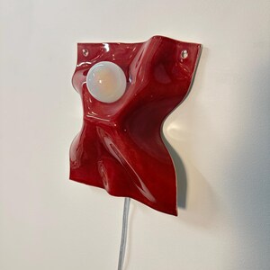 Puede incluir: Una l&aacute;mpara de pared con una base de cer&aacute;mica o pl&aacute;stico de color rojo intenso brillante, de forma irregular con una textura ondulada y plegada. Una bombilla redonda de color blanco esmerilado est&aacute; centrada. Dos peque&ntilde;os agujeros son visibles.