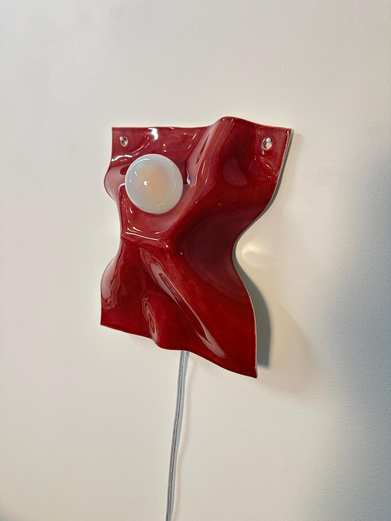 Puede incluir: Una l&aacute;mpara de pared con una base de cer&aacute;mica o pl&aacute;stico de color rojo intenso brillante, de forma irregular con una textura ondulada y plegada. Una bombilla redonda de color blanco esmerilado est&aacute; centrada. Dos peque&ntilde;os agujeros son visibles.