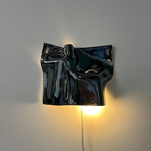 May include: A wall-mounted lamp featuring a glossy, black, undulating surface with a fluid, organic shape. The fixture emits a warm, glowing light from behind its lower edge, casting a soft illumination against the wall and highlighting its curves.