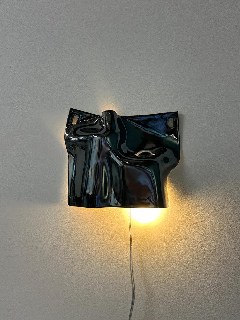May include: A wall-mounted lamp featuring a glossy, black, undulating surface with a fluid, organic shape. The fixture emits a warm, glowing light from behind its lower edge, casting a soft illumination against the wall and highlighting its curves.
