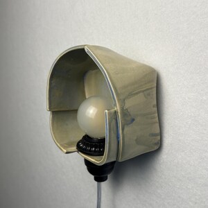 Puede incluir: Un aplique de pared con una pantalla de cer&aacute;mica de color beige claro curvada y bordes con tonos azulados. La l&aacute;mpara incluye una bombilla redonda de color blanco esmerilado en un casquillo negro, con un cable de alimentaci&oacute;n transparente.