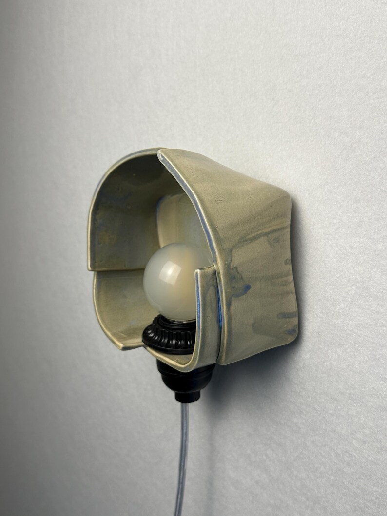Puede incluir: Un aplique de pared con una pantalla de cer&aacute;mica de color beige claro curvada y bordes con tonos azulados. La l&aacute;mpara incluye una bombilla redonda de color blanco esmerilado en un casquillo negro, con un cable de alimentaci&oacute;n transparente.