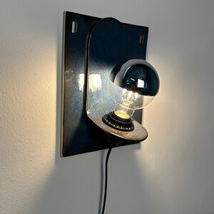 May include: A wall-mounted lamp featuring a dark, rectangular metal backplate with a rounded top section. A circular metal shelf extends from the center, holding a light bulb with a reflective silver top half. A black power cord hangs downward.