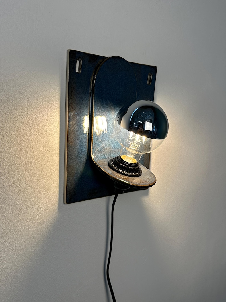 May include: A wall-mounted lamp featuring a dark, rectangular metal backplate with a rounded top section. A circular metal shelf extends from the center, holding a light bulb with a reflective silver top half. A black power cord hangs downward.