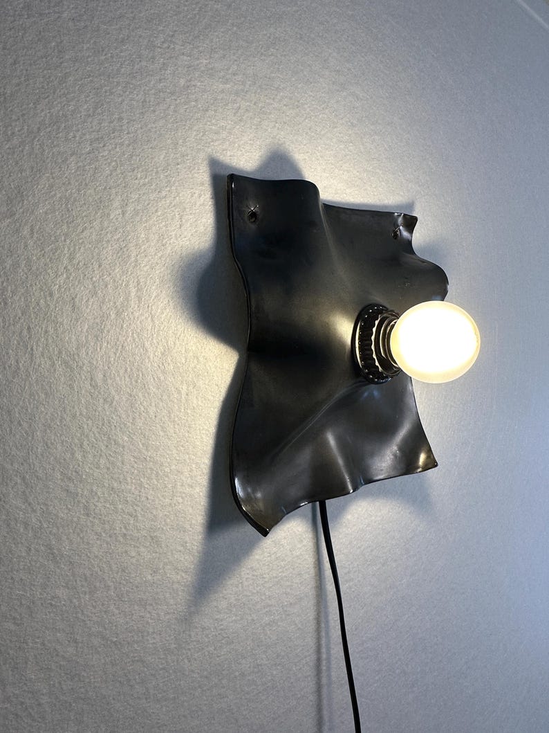 May include: A wall-mounted lamp featuring a matte black, irregularly shaped, wavy metal backplate. A single round, glowing light bulb is mounted on the right side of the plate. A thin black power cord extends downward from the base of the light fixture.