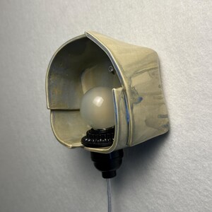 Puede incluir: Un aplique de pared con una pantalla de cer&aacute;mica curva de color claro y sutiles acentos de esmalte azulado. La l&aacute;mpara incluye un casquillo negro que sostiene una bombilla redonda de vidrio esmerilado y un cable fino y transparente.