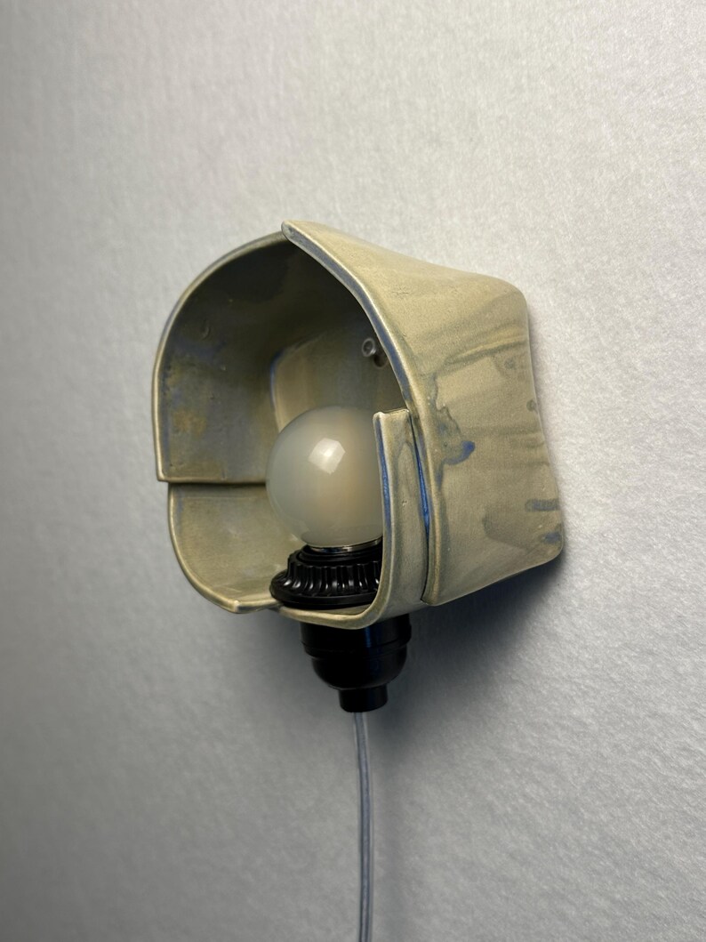 Puede incluir: Un aplique de pared con una pantalla de cer&aacute;mica curva de color claro y sutiles acentos de esmalte azulado. La l&aacute;mpara incluye un casquillo negro que sostiene una bombilla redonda de vidrio esmerilado y un cable fino y transparente.