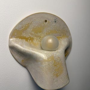 May include: A wall-mounted ceramic sconce featuring a curved, organic shape with a glossy, mottled cream and yellow glaze. The fixture includes a central, spherical white glass bulb that protrudes from the center of the textured ceramic base.