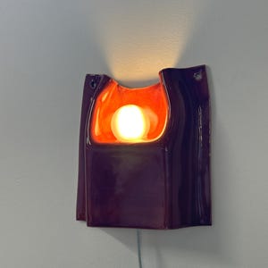 Puede incluir: Un aplique de pared de cer&aacute;mica con un exterior de color morado oscuro brillante y un interior de color naranja intenso. La l&aacute;mpara tiene una forma org&aacute;nica y curva con una abertura central que contiene una bombilla esf&eacute;rica brillante y dos orificios de montaje.