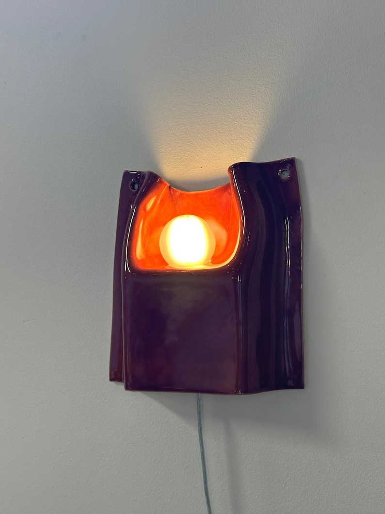 Puede incluir: Un aplique de pared de cer&aacute;mica con un exterior de color morado oscuro brillante y un interior de color naranja intenso. La l&aacute;mpara tiene una forma org&aacute;nica y curva con una abertura central que contiene una bombilla esf&eacute;rica brillante y dos orificios de montaje.