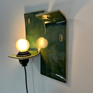 May include: A wall-mounted ceramic sconce featuring a glossy, deep green glaze with a curved, wave-like silhouette. The fixture includes a black socket holding a single, glowing, frosted spherical light bulb, connected by a black electrical cord.