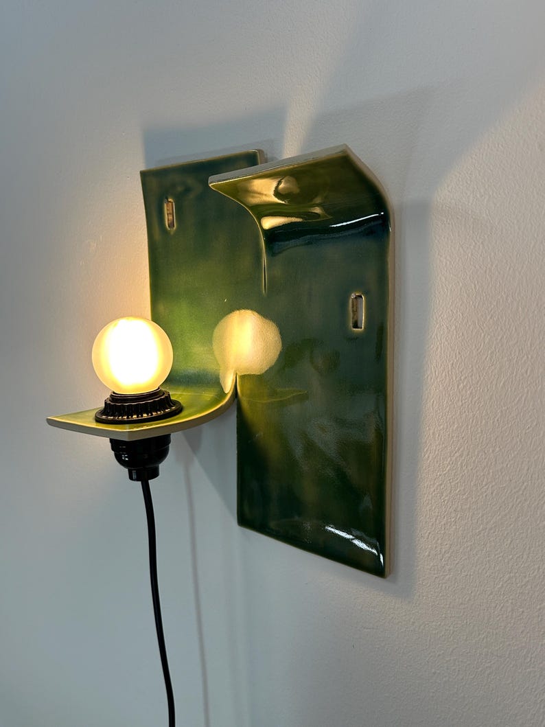 May include: A wall-mounted ceramic sconce featuring a glossy, deep green glaze with a curved, wave-like silhouette. The fixture includes a black socket holding a single, glowing, frosted spherical light bulb, connected by a black electrical cord.