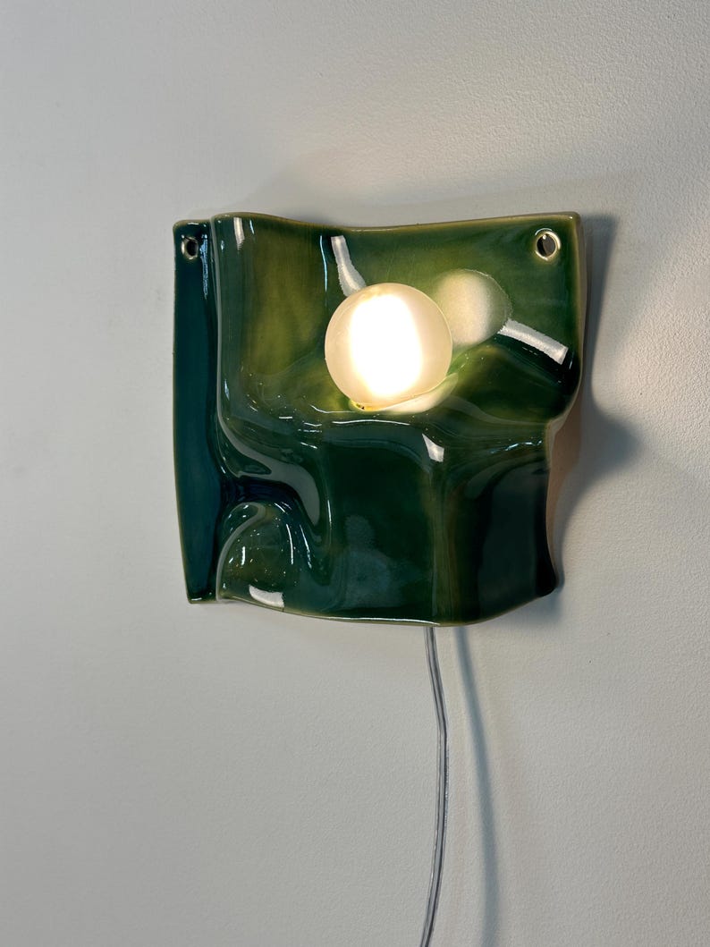 May include: A wall-mounted ceramic light fixture with a glossy, deep green, undulating surface. The piece features a circular, glowing white bulb integrated into the center of the curved form, with two small holes at the top corners for mounting.