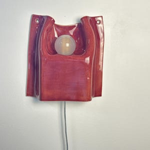 Puede incluir: Un aplique de pared con una carcasa de cer&aacute;mica roja oscura brillante de forma escultural y plegada. La l&aacute;mpara incluye una bombilla central esf&eacute;rica de vidrio esmerilado y dos peque&ntilde;os orificios de montaje circulares en las esquinas superiores.