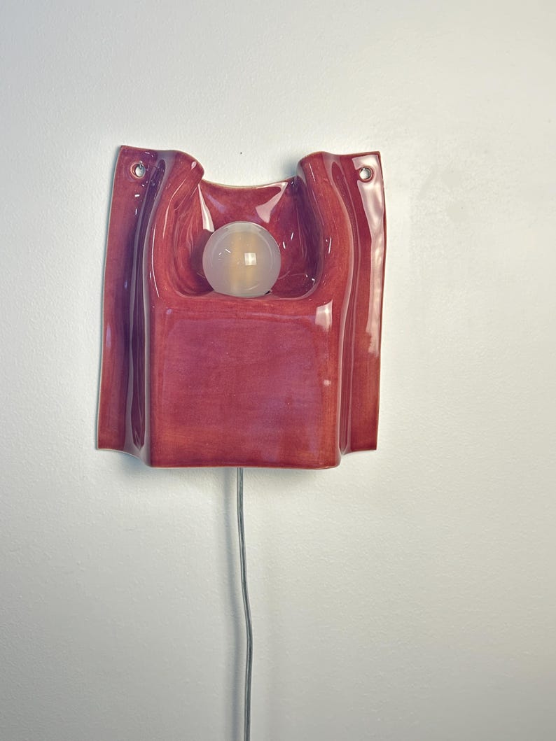 Puede incluir: Un aplique de pared con una carcasa de cer&aacute;mica roja oscura brillante de forma escultural y plegada. La l&aacute;mpara incluye una bombilla central esf&eacute;rica de vidrio esmerilado y dos peque&ntilde;os orificios de montaje circulares en las esquinas superiores.