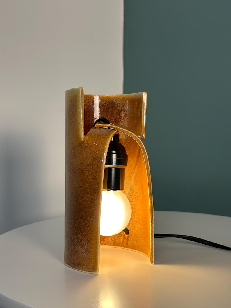 Puede incluir: Una l&aacute;mpara de mesa cil&iacute;ndrica de cer&aacute;mica con un esmalte brillante de color &aacute;mbar marr&oacute;n moteado. La estructura presenta un dise&ntilde;o curvo con frente abierto que revela un casquillo negro con una bombilla blanca encendida y un cable negro.