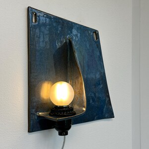 May include: A modern wall sconce with a square, blue-toned metal shade. The shade has a unique folded design and a warm, glowing light bulb. The fixture is mounted on a white wall, showcasing its contemporary aesthetic.