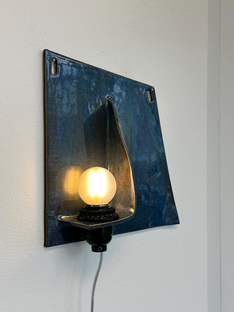 May include: A modern wall sconce with a square, blue-toned metal shade. The shade has a unique folded design and a warm, glowing light bulb. The fixture is mounted on a white wall, showcasing its contemporary aesthetic.
