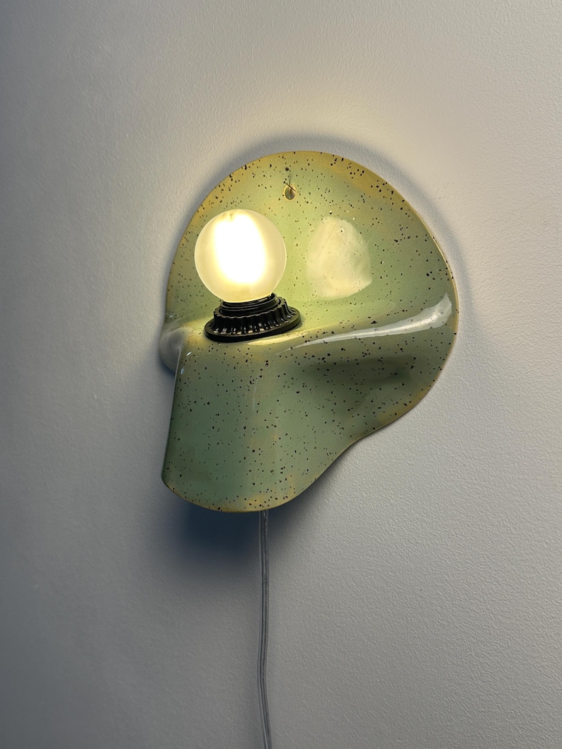 Puede incluir: Un aplique de pared con una pantalla de cer&aacute;mica curva de color verde p&aacute;lido con un patr&oacute;n de motas oscuras. La l&aacute;mpara incluye un casquillo circular negro que sostiene una bombilla esf&eacute;rica esmerilada encendida y un cable blanco fino.