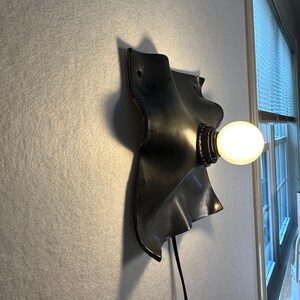 May include: A wall-mounted lamp featuring a matte black, wavy, organic-shaped metal backplate. A single round, glowing white light bulb is attached to the center of the plate. A thin black power cord extends downward from the base of the fixture.