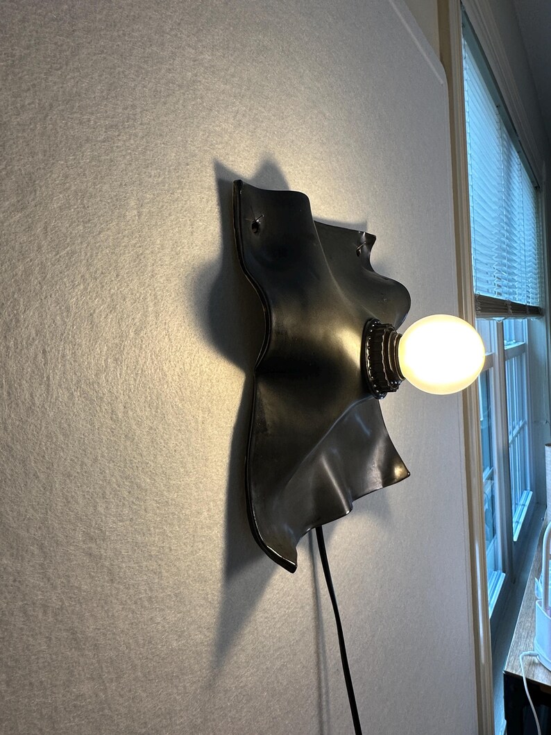 May include: A wall-mounted lamp featuring a matte black, wavy, organic-shaped metal backplate. A single round, glowing white light bulb is attached to the center of the plate. A thin black power cord extends downward from the base of the fixture.