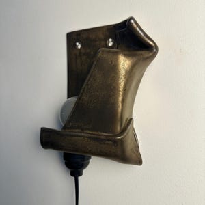 May include: A wall-mounted light fixture featuring a dark, metallic, bronze-toned shade with a folded, sculptural form. The fixture includes a black socket, a white light bulb, and a black power cord extending downward from the base.
