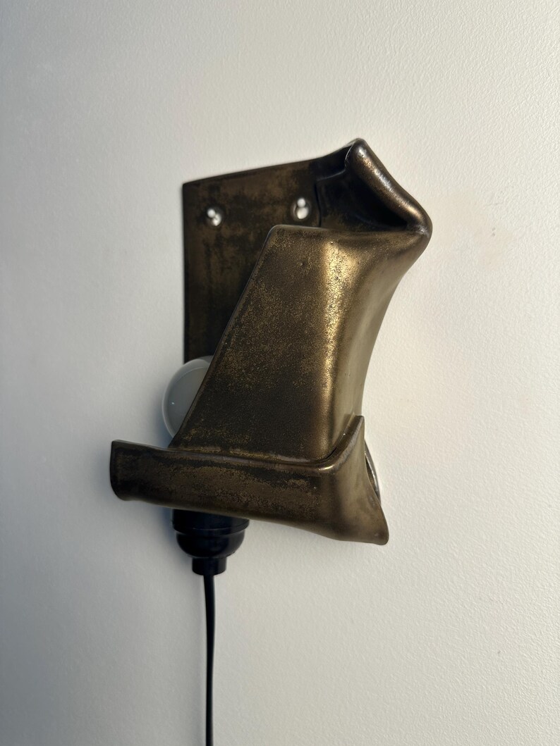 May include: A wall-mounted light fixture featuring a dark, metallic, bronze-toned shade with a folded, sculptural form. The fixture includes a black socket, a white light bulb, and a black power cord extending downward from the base.