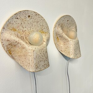 May include: Two wall-mounted ceramic sconces with a speckled cream and brown finish. Each fixture features a curved, organic shape with a central circular opening holding a small, round, frosted white light bulb and a thin, clear power cord.