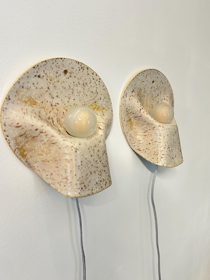 May include: Two wall-mounted ceramic sconces with a speckled cream and brown finish. Each fixture features a curved, organic shape with a central circular opening holding a small, round, frosted white light bulb and a thin, clear power cord.