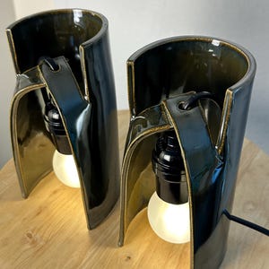 Puede incluir: Dos l&aacute;mparas de mesa cil&iacute;ndricas de cer&aacute;mica con un esmalte brillante de color verde oliva oscuro. Cada l&aacute;mpara presenta una estructura curva de lados abiertos con un casquillo negro y una bombilla blanca brillante suspendida de un brazo.