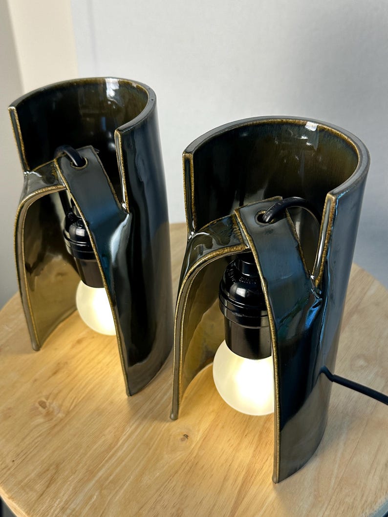 Puede incluir: Dos l&aacute;mparas de mesa cil&iacute;ndricas de cer&aacute;mica con un esmalte brillante de color verde oliva oscuro. Cada l&aacute;mpara presenta una estructura curva de lados abiertos con un casquillo negro y una bombilla blanca brillante suspendida de un brazo.