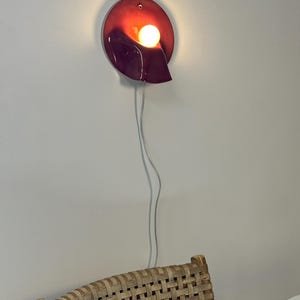 May include: A wall-mounted lamp featuring a glossy, deep red, oval-shaped ceramic base with a curved, sculptural form. A single, warm-glowing spherical light bulb is nestled within the centre of the fixture, connected by a thin, clear power cord.