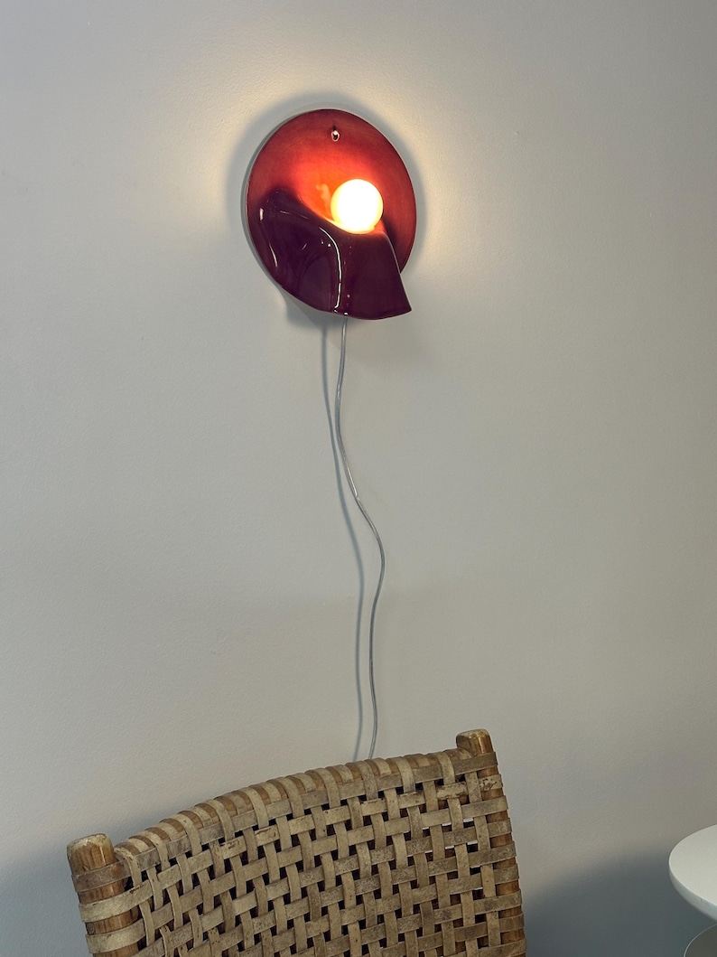 May include: A wall-mounted lamp featuring a glossy, deep red, oval-shaped ceramic base with a curved, sculptural form. A single, warm-glowing spherical light bulb is nestled within the centre of the fixture, connected by a thin, clear power cord.