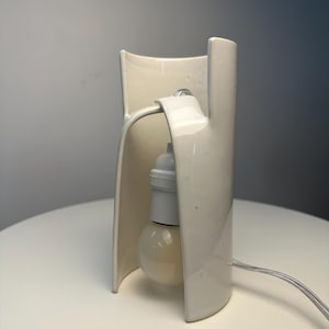 May include: A cream-colored ceramic table lamp with a curved, open-sided structure. The lamp features a central white socket holding a round light bulb, with a clear electrical cord extending from the side of the smooth, glossy ceramic base.