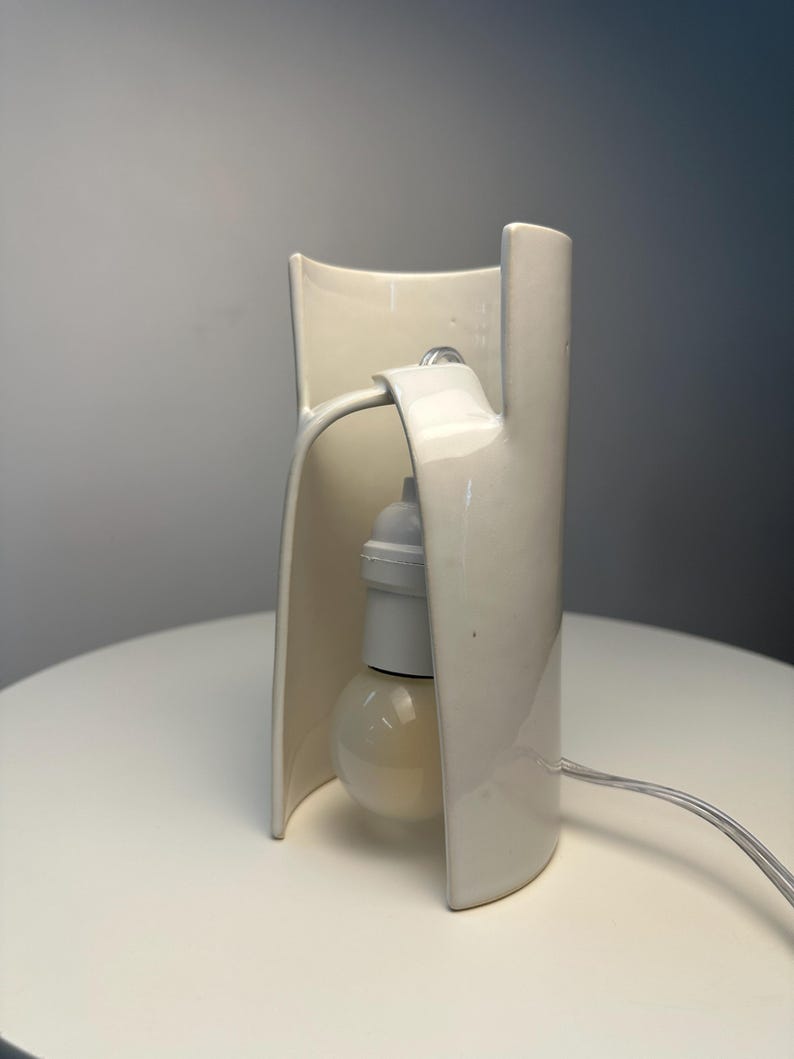 May include: A cream-colored ceramic table lamp with a curved, open-sided structure. The lamp features a central white socket holding a round light bulb, with a clear electrical cord extending from the side of the smooth, glossy ceramic base.