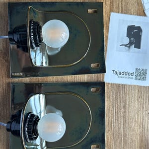 May include: Two ceramic wall sconces with a dark teal glaze and a gold trim. Each sconce features a black bulb socket and a round, frosted light bulb. The sconces are rectangular with a curved top. A paper with the text "Tajaddod" is on the right.