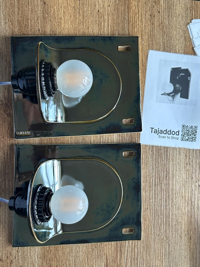 May include: Two ceramic wall sconces with a dark teal glaze and a gold trim. Each sconce features a black bulb socket and a round, frosted light bulb. The sconces are rectangular with a curved top. A paper with the text "Tajaddod" is on the right.