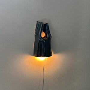 May include: A dark, glossy ceramic wall light with an irregular, organic shape. The fixture features a central cutout and a downward-facing opening that emits a warm, glowing light. A thin, light-coloured power cord extends from the bottom of the lamp.
