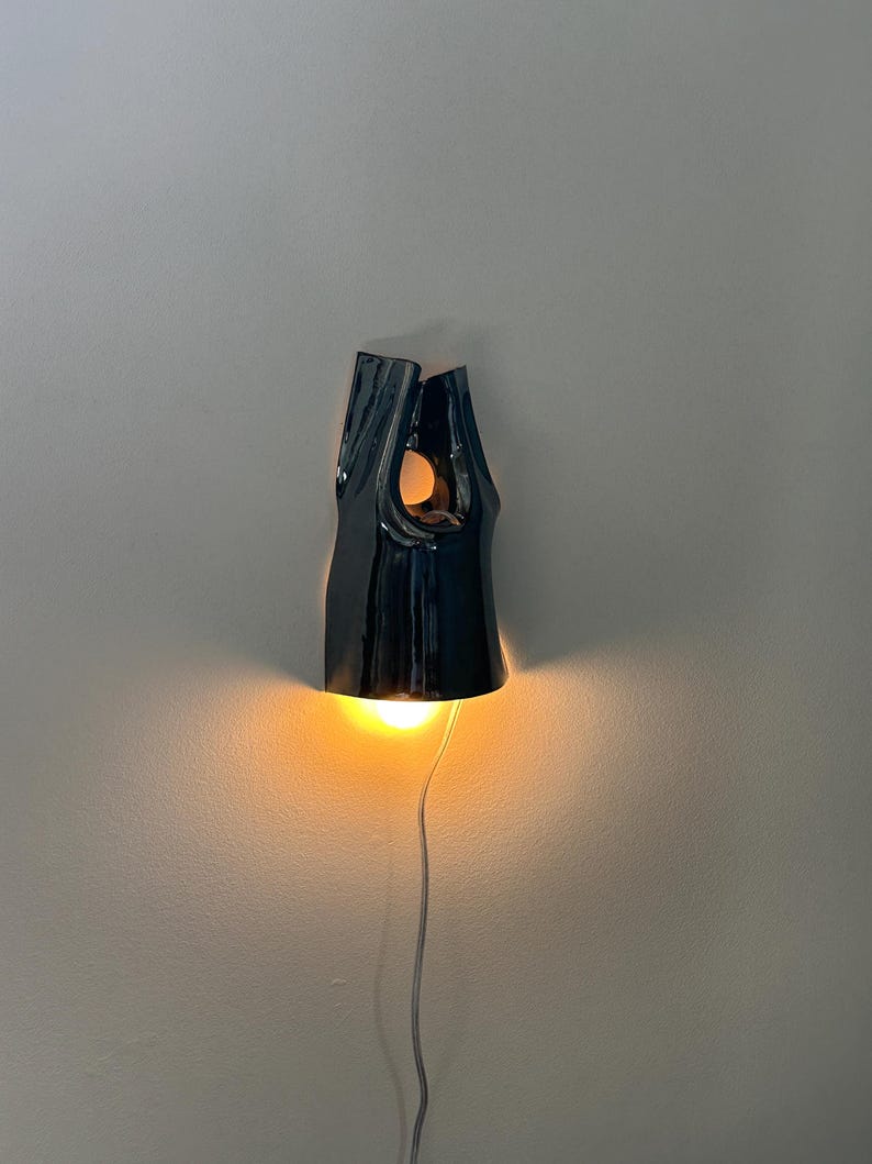 May include: A dark, glossy ceramic wall light with an irregular, organic shape. The fixture features a central cutout and a downward-facing opening that emits a warm, glowing light. A thin, light-coloured power cord extends from the bottom of the lamp.