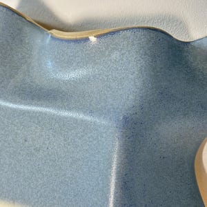 May include: A ceramic bowl with a wavy, undulating rim and a speckled light blue glaze. The surface features a textured finish with darker blue flecks throughout, and the edges are highlighted with a slightly deeper blue tone.