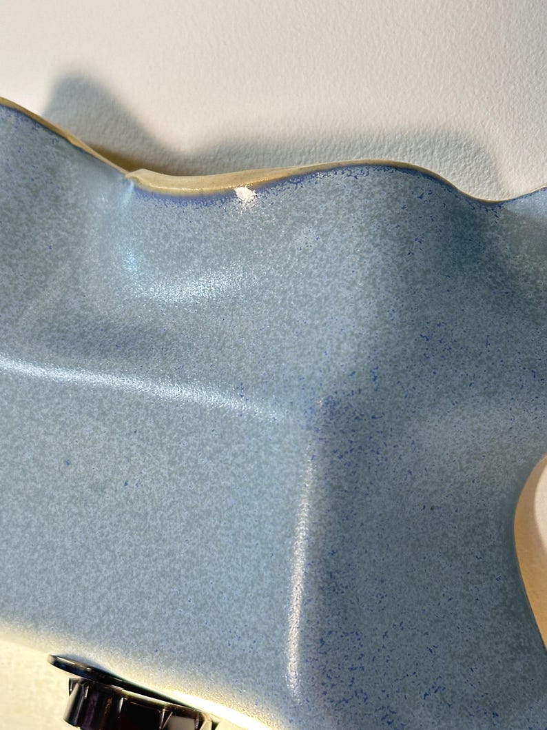 May include: A ceramic bowl with a wavy, undulating rim and a speckled light blue glaze. The surface features a textured finish with darker blue flecks throughout, and the edges are highlighted with a slightly deeper blue tone.