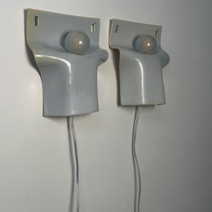 May include: Two wall-mounted light fixtures featuring a smooth, light gray ceramic body with a curved, folded silhouette. Each fixture includes a single round, frosted glass bulb centered on the upper portion and a clear electrical cord extending downward.