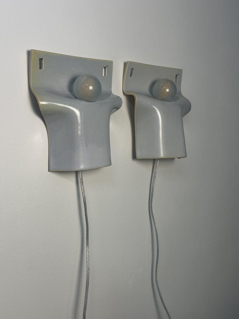 May include: Two wall-mounted light fixtures featuring a smooth, light gray ceramic body with a curved, folded silhouette. Each fixture includes a single round, frosted glass bulb centered on the upper portion and a clear electrical cord extending downward.