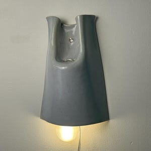 K&ouml;nnte beinhalten: Eine Wandleuchte mit einem glatten, gl&auml;nzenden, hellgrauen Keramikschirm in einer ausgestellten, glockenf&ouml;rmigen Silhouette. Die Leuchte hat eine zentrale &Ouml;ffnung an der Oberseite und strahlt ein warmes, sanftes Licht von einer runden Gl&uuml;hbirne aus.