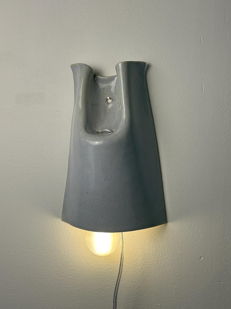 K&ouml;nnte beinhalten: Eine Wandleuchte mit einem glatten, gl&auml;nzenden, hellgrauen Keramikschirm in einer ausgestellten, glockenf&ouml;rmigen Silhouette. Die Leuchte hat eine zentrale &Ouml;ffnung an der Oberseite und strahlt ein warmes, sanftes Licht von einer runden Gl&uuml;hbirne aus.