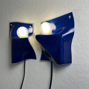May include: Two wall-mounted lamps featuring glossy, deep blue ceramic shades with irregular, folded forms. Each lamp includes a single, spherical, glowing white light bulb nestled within the ceramic structure and a black power cord extending downward.